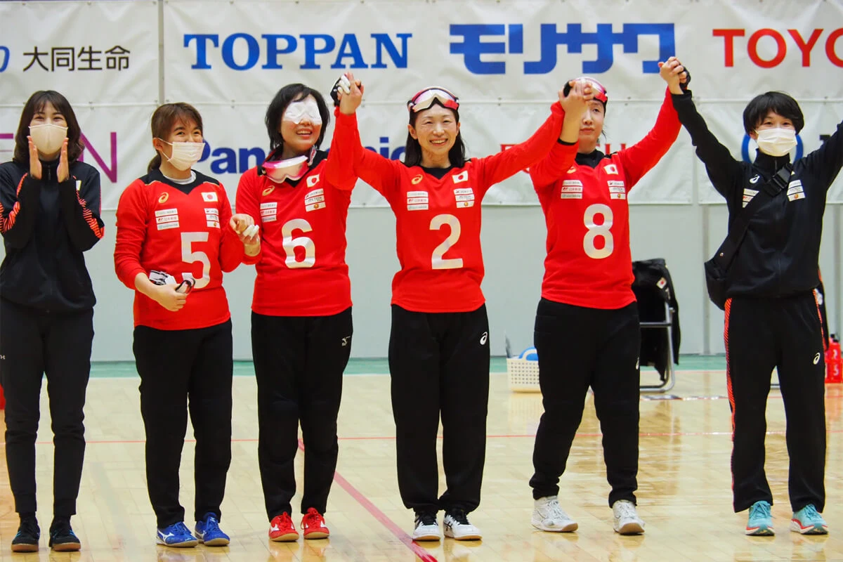 写真:2021年12月のジャパンパラで、赤いユニフォームを着たゴールボール選手4人が手をつないで笑顔を見せる様子。中央が浦田理恵選手